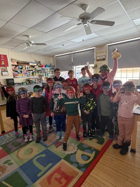 Group of elementary students standing together on a classroom rug wearing colorful safety goggles, posing and smiling during a science activity.
