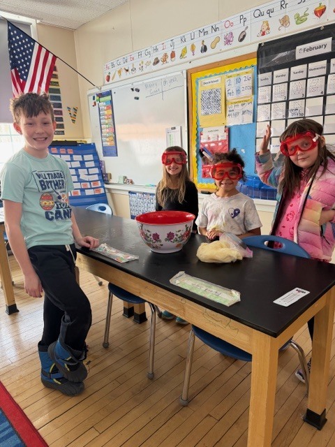 Four students wearing safety goggles stand around a lab table with a red bowl of ice and experiment supplies, smiling at the camera.