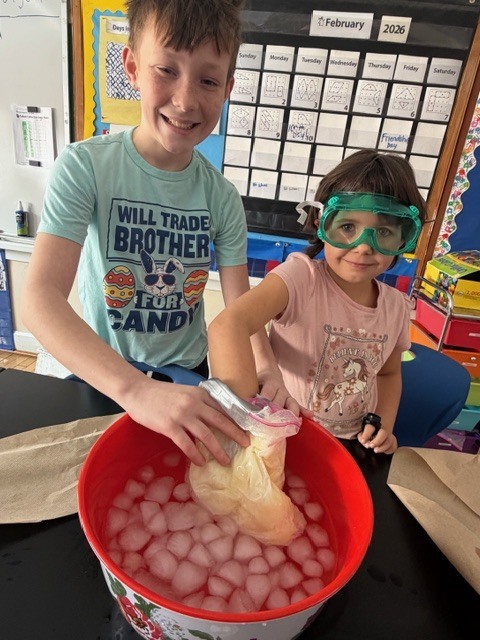 Two students wearing safety goggles place a sealed plastic bag into a large red bowl filled with ice, conducting a classroom science experiment.