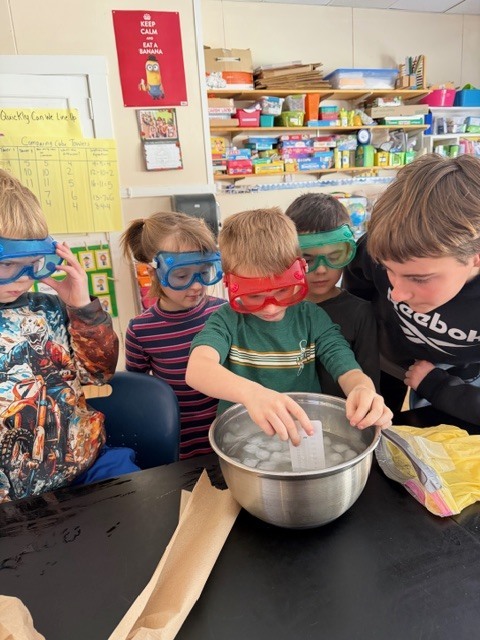 Five students in goggles gather around a metal bowl of ice while one student dips a measuring strip or paper into the water as others watch.
