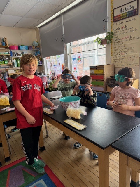 Four students around a lab table with goggles and materials prepare for an ice-bath science activity in a classroom.