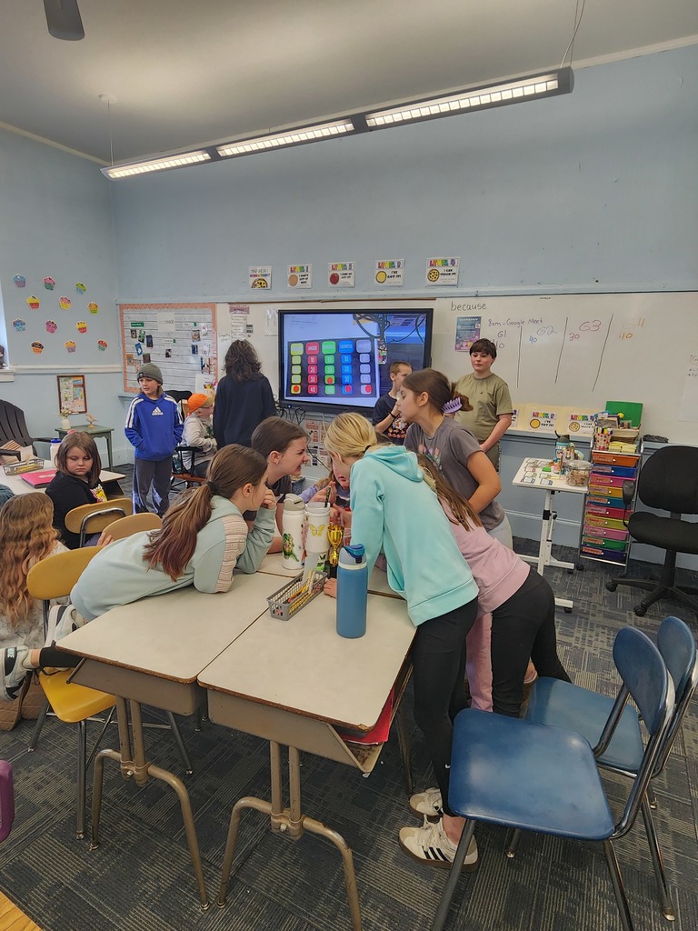 Students gather around desks in a classroom, leaning in and talking together during a group activity, with an interactive board visible in the background.