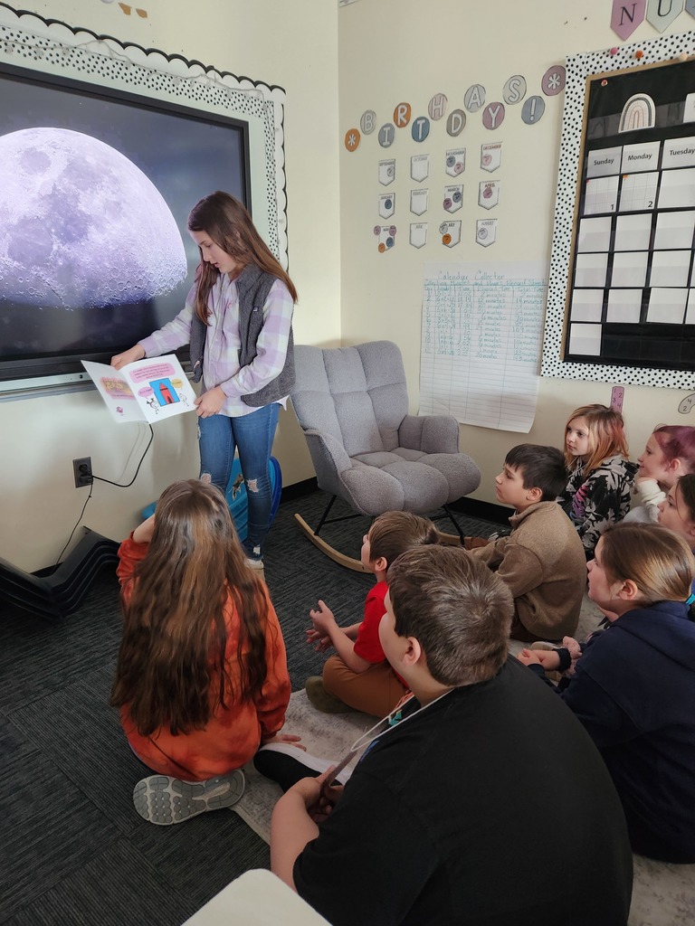 Student stands at the front of a classroom reading from a picture book beside a large screen showing a close-up image of the moon, while several elementary students sit on the floor listening.