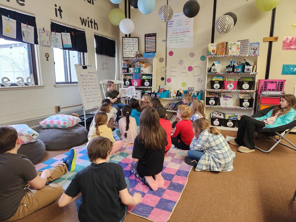 A group of elementary students sit on a colorful rug in a classroom library area while a student reads aloud from a book near a whiteboard and shelves labeled “READ.”