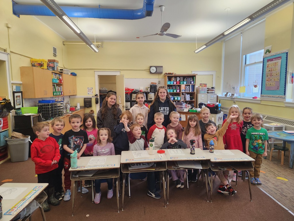 Large group of kindergarten-aged students and a few older student helpers pose together behind classroom desks, smiling at the camera.