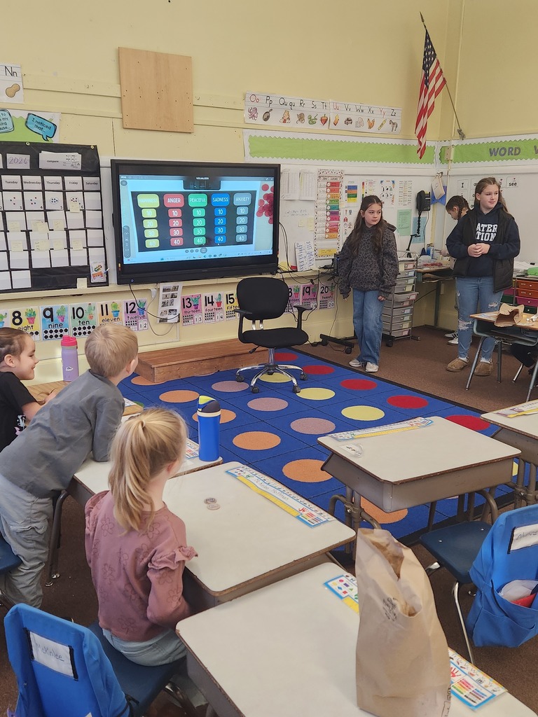 Older students stand at the front of an elementary classroom near an interactive board displaying a feelings-style quiz game, while younger students sit at desks watching.