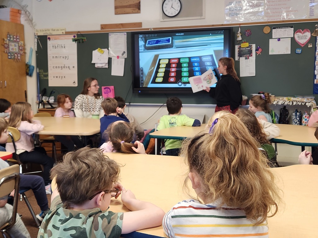 Students sit around tables facing a classroom screen showing a feelings-themed quiz board, while a student at the front holds a book and presents to the class.