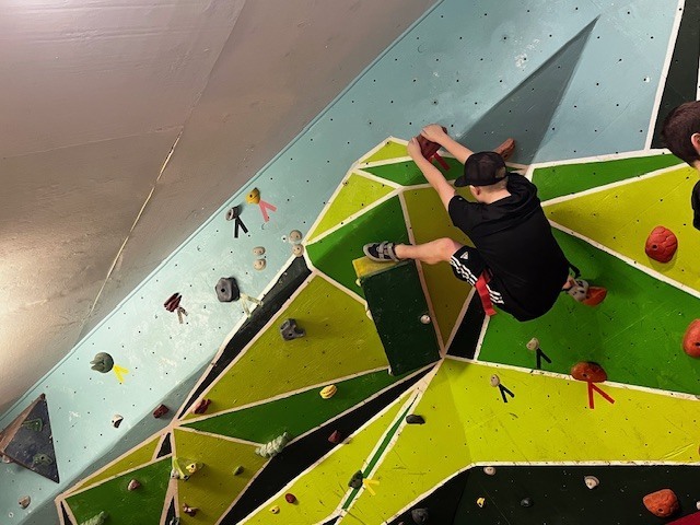 Student bouldering on an angled green climbing wall, reaching for holds while secured with a harness.