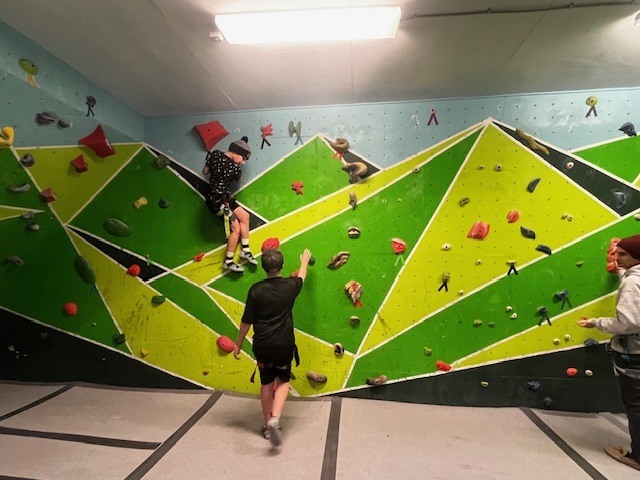 Wide view of multiple students climbing and spotting on a green indoor bouldering wall with geometric panels.