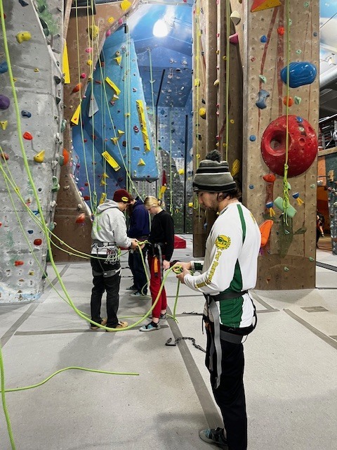 Students in harnesses preparing ropes and belay equipment on the floor of an indoor climbing gym.