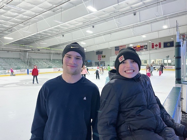 Two students smiling at the camera while standing beside the boards at an indoor ice rink with skaters behind them.