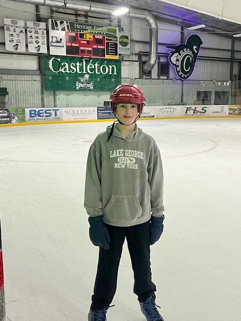 Student wearing a red helmet and winter gloves standing on the ice rink posing for a photo near the goal area.