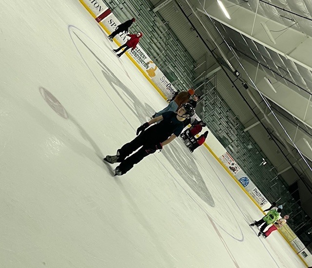 Student ice skating at an indoor rink with others in the background, wearing a helmet and winter gear.