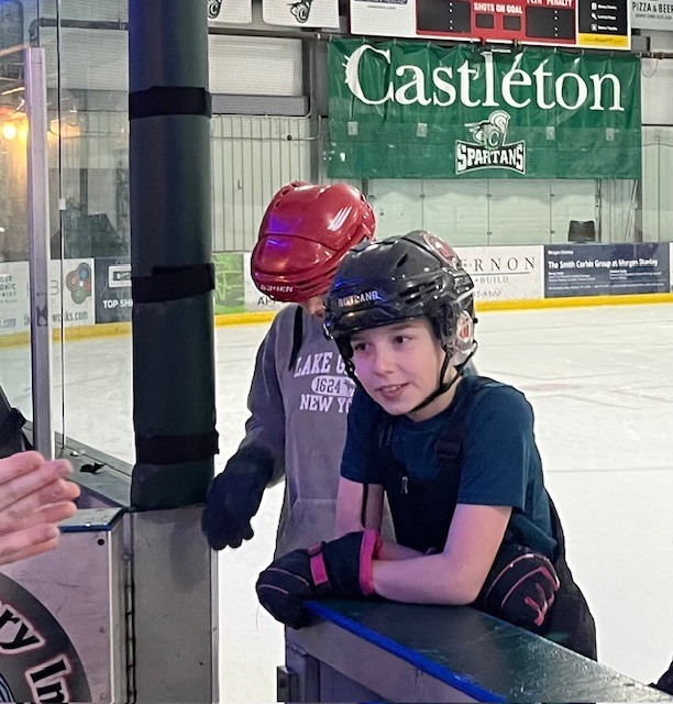 Two helmeted students at the edge of the ice rink listening to an adult just off camera, with a Castleton Spartans banner behind them.