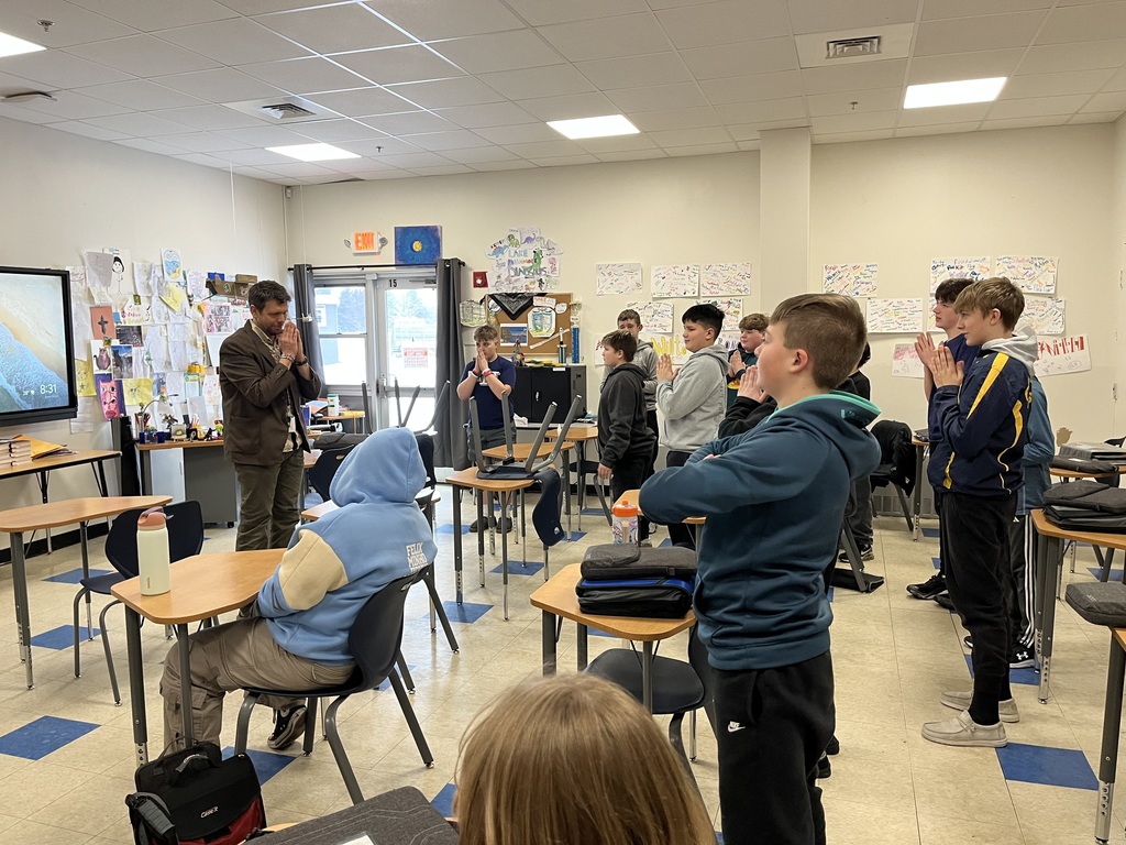 A teacher and group of middle school students stand in a circle around desks with hands together at their chests, practicing a quiet breathing or centering exercise in the classroom.