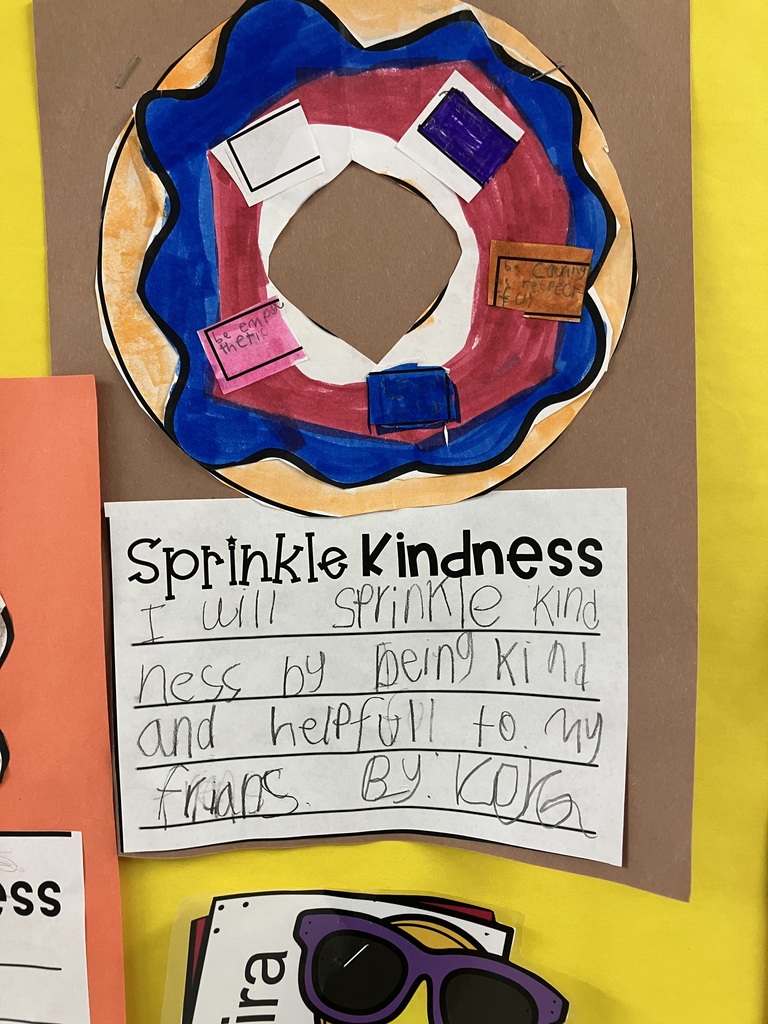  A close-up of a student project on a brown background. The donut features bright blue and pink frosting with small paper sprinkles. The writing on the "Sprinkle Kindness" sheet below the donut is written in large, clear print: "I will sprinkle kindness by being kind and helpfull to my frians. 