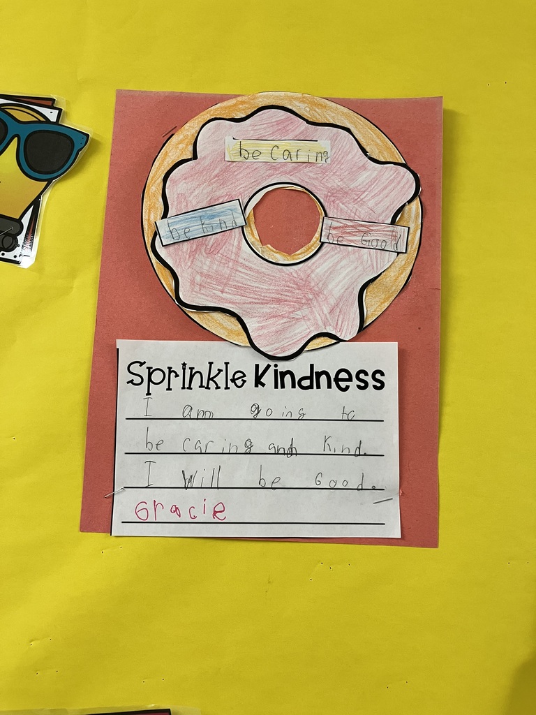 "Sprinkle Kindness" Project A child’s craft project on red construction paper featuring a hand-drawn donut with pink frosting and a brown base. Three small paper "sprinkles" are glued on, with "be caring," "be kind," and "be good" written in pencil. Below the donut, a worksheet titled "Sprinkle Kindness" shows a child’s handwriting in pencil and red marker that reads: "I am going to be caring and kind. I will be good. 