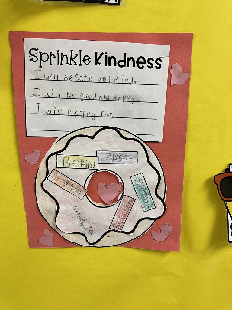A Colorful Kindness Donut A close-up of a student's "Sprinkle Kindness" project. The donut has white frosting with several colorful paper sprinkles glued to it, containing words like "Be Kind," "Be Safe," "Be Good," and "Love." Pink paper hearts are scattered around the donut on a red background. The writing on the worksheet above reads: "I will be safe and kind. I will be good and happy. I will be joyful."  