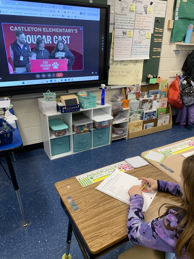 A classroom smartboard displays the CES Cougar Cast morning announcements broadcast while a student sits at a desk watching and working on a worksheet.