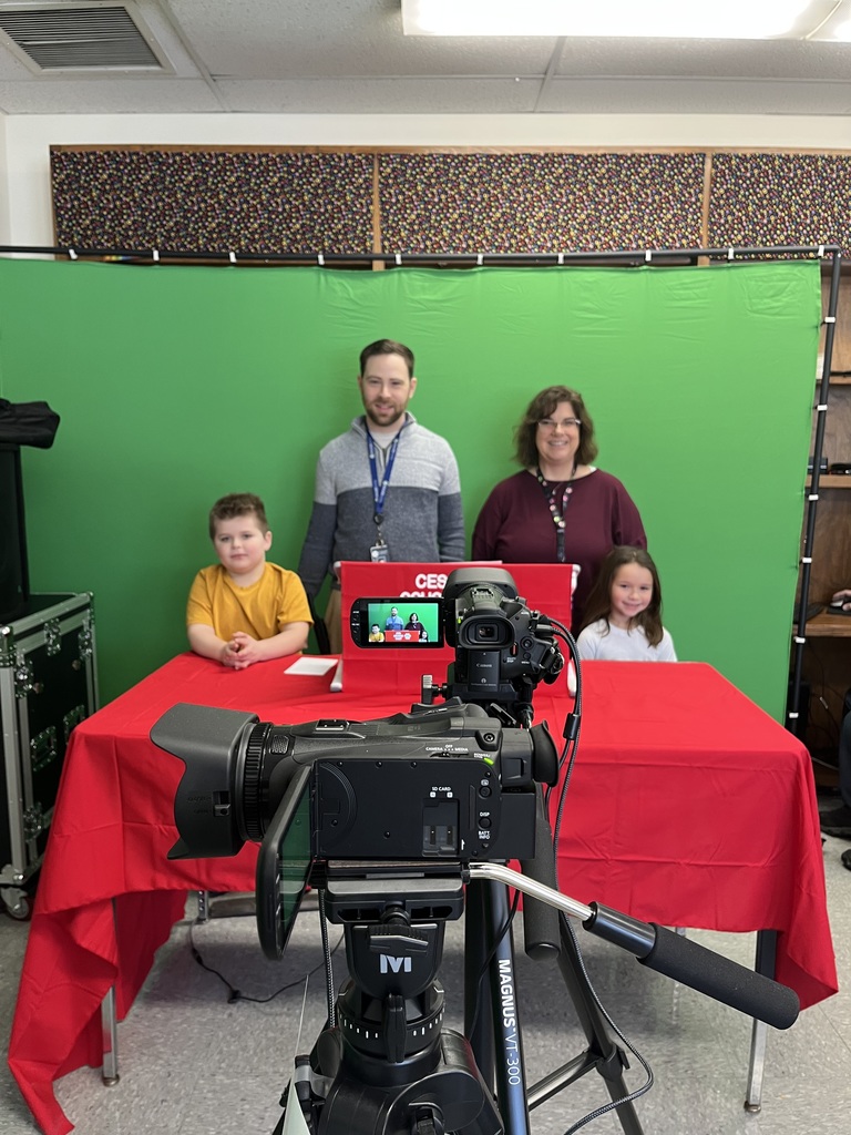 Two staff members and two elementary students sit behind a red desk in front of a green screen while a video camera on a tripod records their school news broadcast.