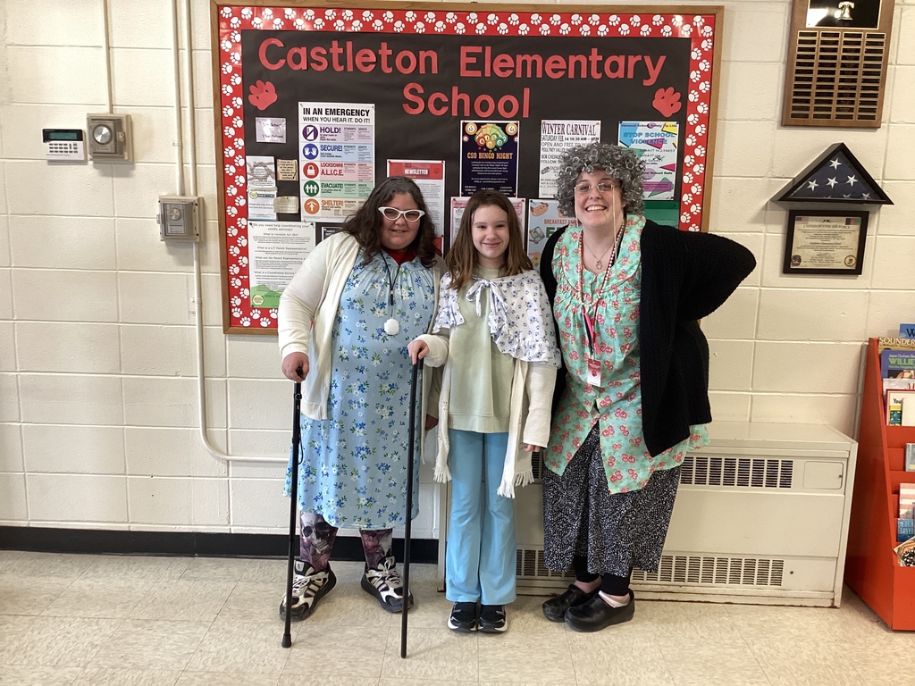 Three people at Castleton Elementary School pose in front of a school bulletin board, dressed like 100-year-olds for the 100th day of school. Two students and a teacher wear gray wigs, layered clothing, and hold canes, smiling for the photo.