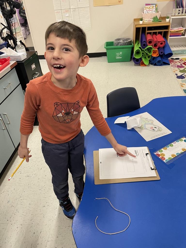 : A smiling boy in an orange shirt with a geometric bear design stands at a blue table. He proudly points to the "Longer Than" section of his worksheet. A piece of tan string is coiled on the table in front of him.  