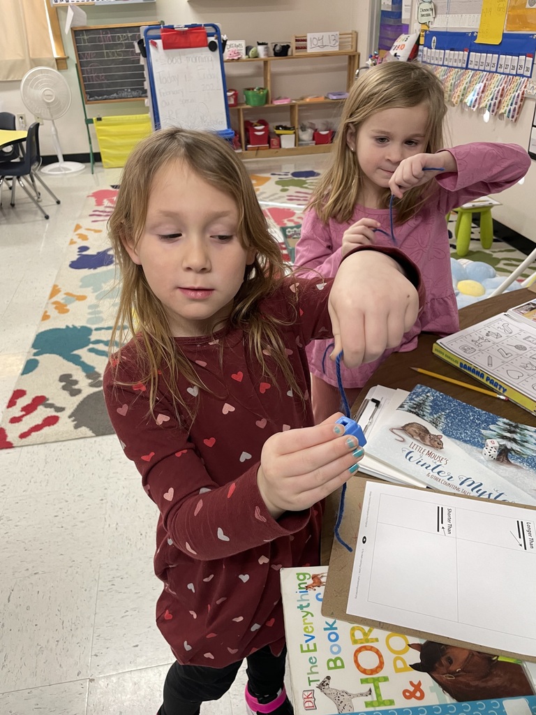  Two young girls focus intently on their task. One girl in the foreground wears a maroon heart-patterned dress, holding a blue string against a small blue plastic cube. Behind her, another girl in a pink shirt holds up her own blue string. 