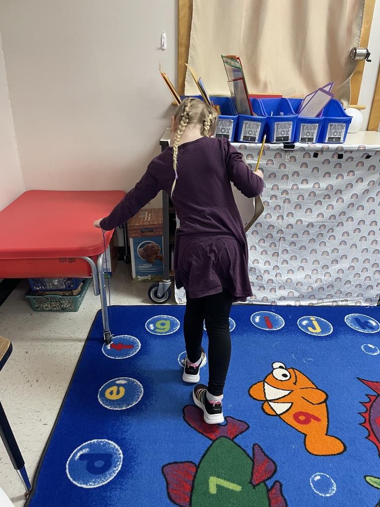  A girl with blonde hair in two French braids and a purple dress stands on a blue alphabet rug. She is leaning over to measure the side of a red sensory table with her string while holding a clipboard and pencil in her other hand.  IMG_5430.jpg: A young boy in a teal "Transformers" shirt stands in a classroom kitchen area. He is holding a pencil in one hand and carefully manipulating a piece of tan string with the other, appearing to compare the two lengths.