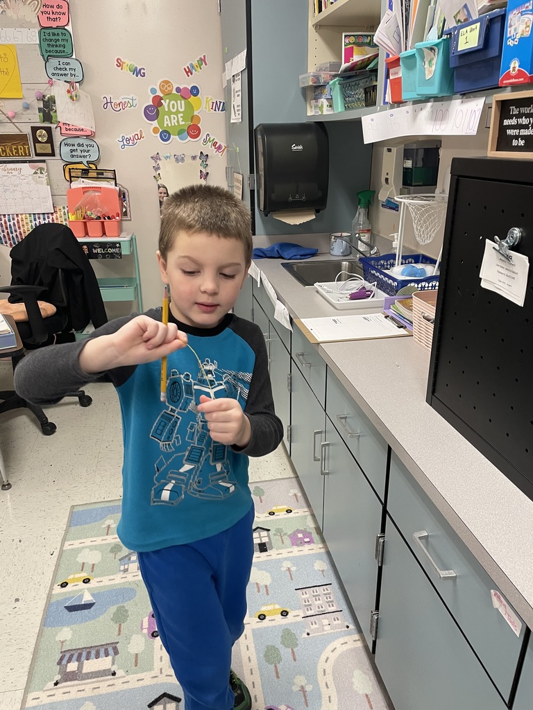 A young boy in a teal "Transformers" shirt stands in a classroom kitchen area. He is holding a pencil in one hand and carefully manipulating a piece of tan string with the other, appearing to compare the two lengths.