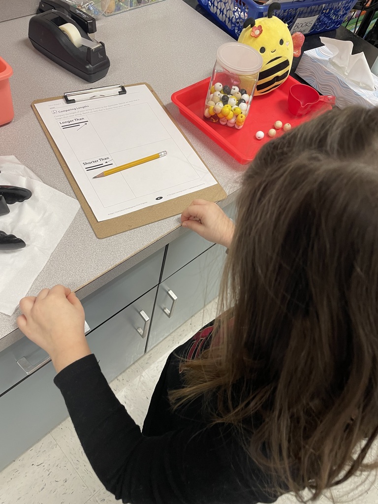 A close-up, over-the-shoulder view of a student stretching a tan string along the edge of a grey countertop to measure its length. A worksheet on a clipboard nearby shows a pencil placed in the "Shorter Than" category.  
