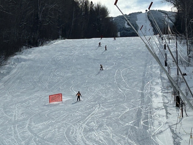 Wide view of a ski slope with several skiers descending at different distances, lift poles and cables running up the right side, and wooded hills beyond.