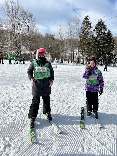 Two elementary students wearing helmets and green bibs stand on skis facing the camera on a sunny slope with other skiers behind them.