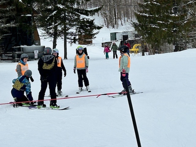 A small group of students in helmets and orange bibs gather around an instructor on skis near a boundary rope at the ski area.