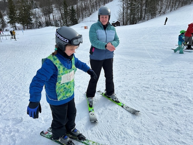An instructor on skis speaks with a young student in a helmet and green bib, both standing on a gentle slope during a lesson.