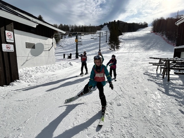 A small group of students on skis glide across a flat area near the base of a ski hill with lift towers and buildings in the background.