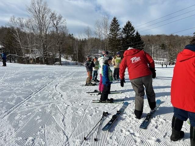 Students in helmets and ski gear stand in a line on a groomed ski slope while an instructor in a red jacket on skis gives directions.