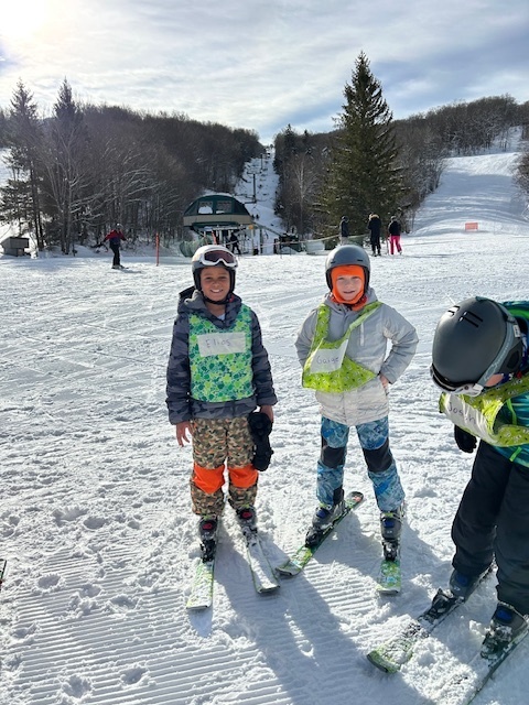 Three young skiers in helmets and numbered bibs stand together on skis near the base area with a chairlift and trail in the background.