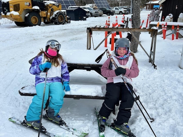 Two young skiers sit on a snow-covered picnic table holding their skis and poles, resting near equipment, cones, and a plowed area.