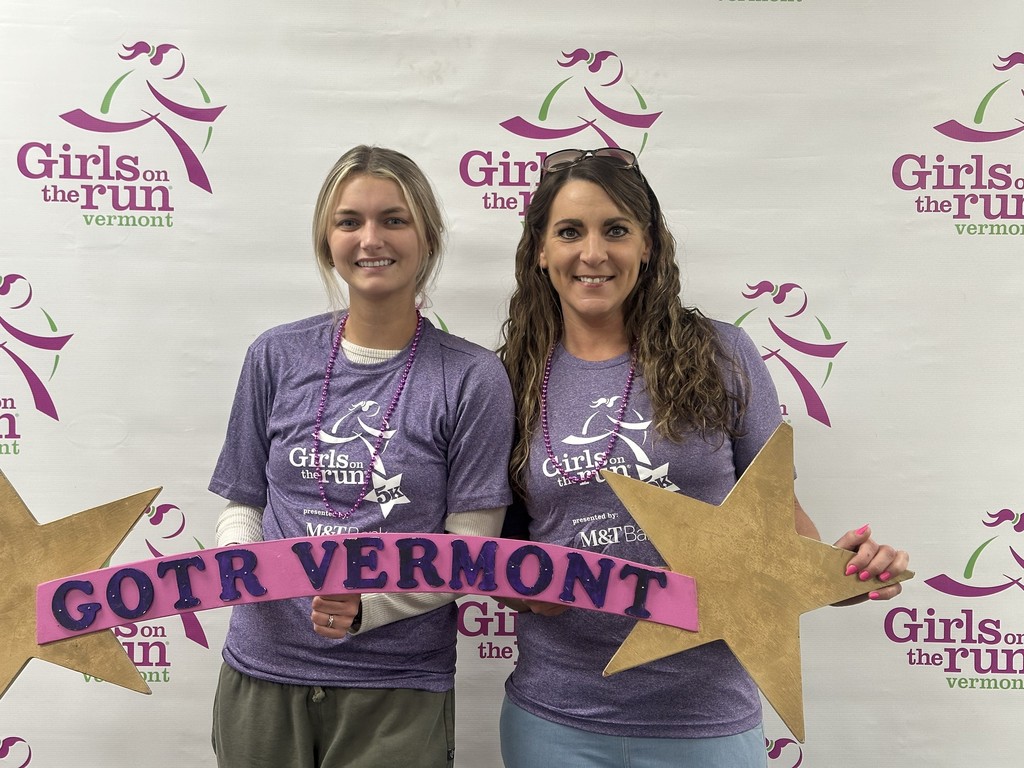 Two adult coaches wearing purple Girls on the Run shirts stand smiling in front of a Girls on the Run Vermont backdrop, holding a pink and gold sign that reads “GOTR Vermont.”