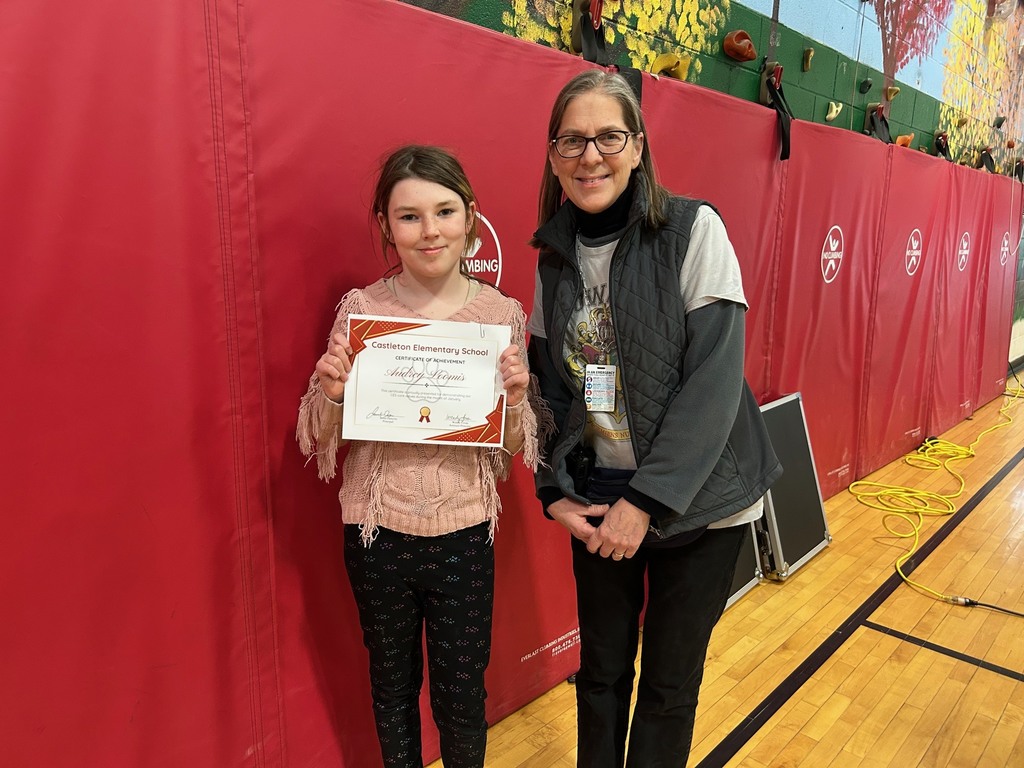 An elementary student holds a certificate next to a staff member in a school gym with red wall padding in the background.