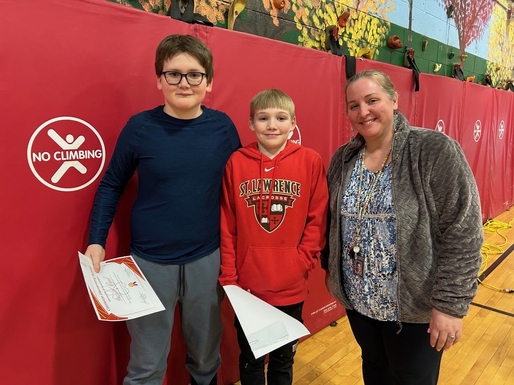 Two elementary students stand with a staff member in the gym, holding certificates and smiling after an awards presentation.