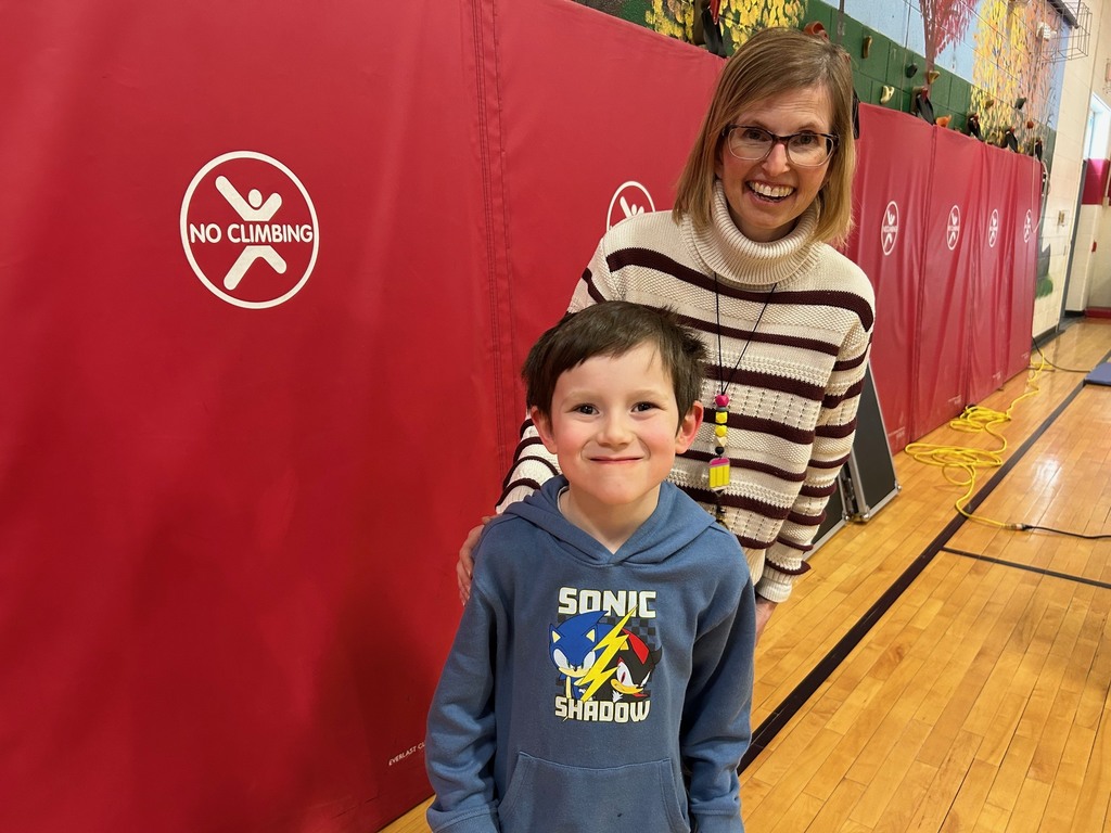 An elementary student smiles in front of a red gym wall while a teacher stands behind with a hand on the student’s shoulder.