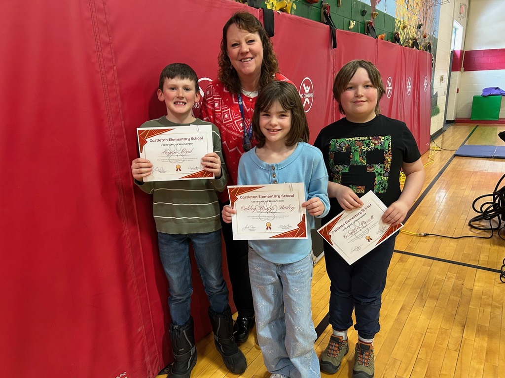 Three elementary students holding achievement certificates stand with a teacher in front of red gym wall padding.