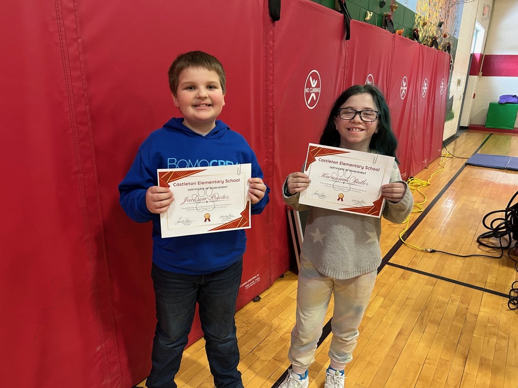 Two elementary students hold Castleton Elementary School certificates while standing in front of red gym wall padding.