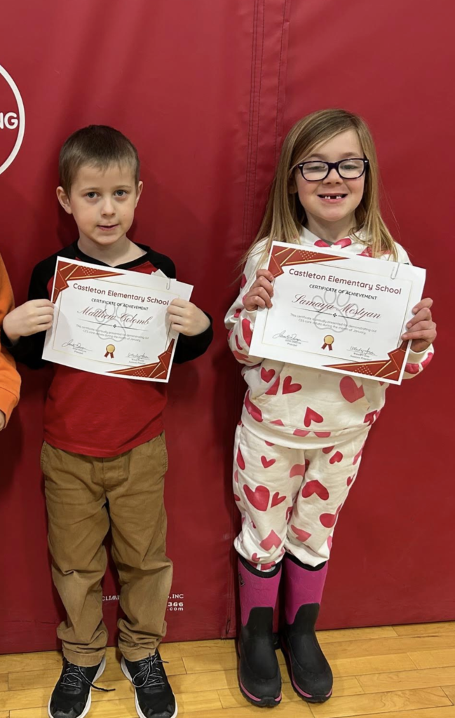 Two elementary students stand in front of a red gym wall holding Castleton Elementary School certificates and smiling at the camera.