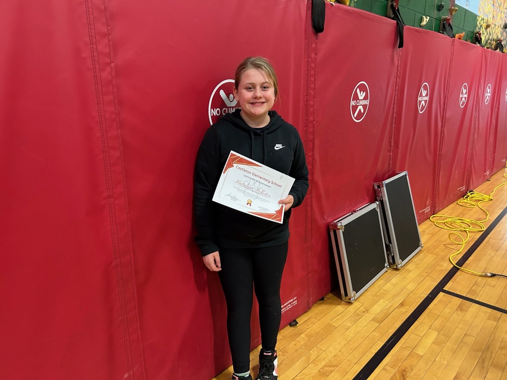 A student dressed in black athletic clothing stands against red gym wall pads holding a Castleton Elementary School certificate of achievement.