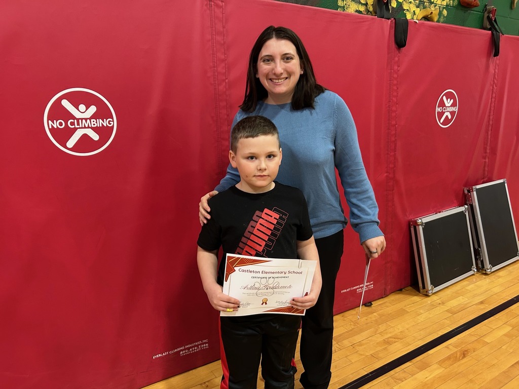 An adult and a student stand side by side in a gym, each holding a Castleton Elementary School certificate of achievement, smiling toward the camera.