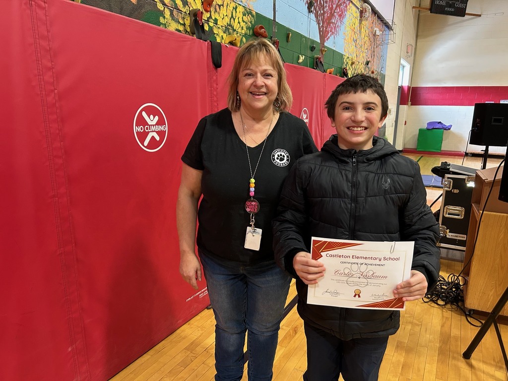 A smiling student holds a Castleton Elementary School certificate while standing next to a staff member in a gym with red wall padding and gym equipment visible in the background.