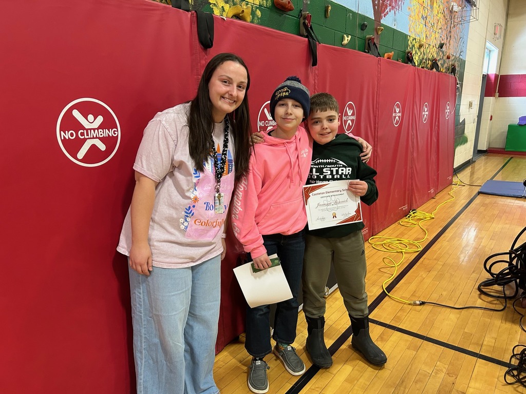 Three people stand in a school gym in front of red wall pads with “No Climbing” signs. Two students stand with an adult staff member, and one student holds a Castleton Elementary School certificate of achievement.