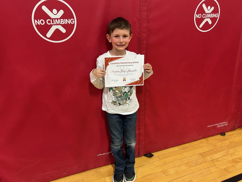 A student stands in front of red gym wall pads holding a Castleton Elementary School certificate of achievement and smiling.